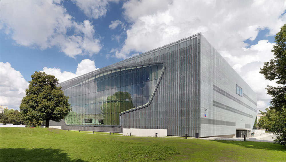 Modern angular building with a perforated metal facade and large curved glass window, set beside green lawn and trees under a clear sky