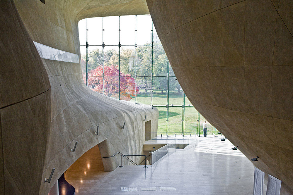 Modern museum interior with curving beige walls, a large glass window overlooking a park with autumn foliage, and a person walking through the gallery