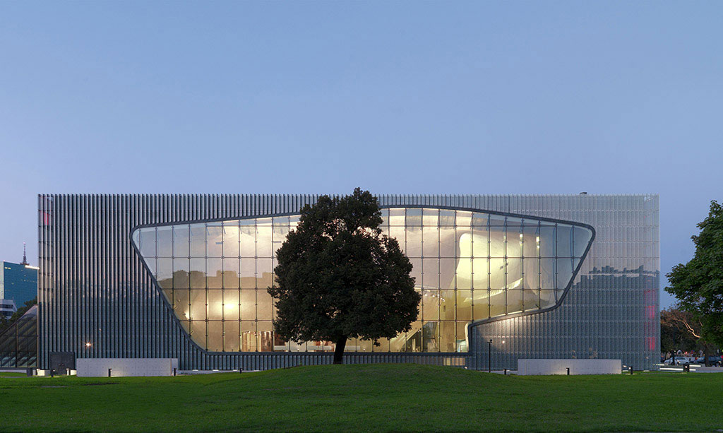 A modern glass and metal building with a curved illuminated opening, a large tree in front on a grassy lawn at dusk, moody and warm