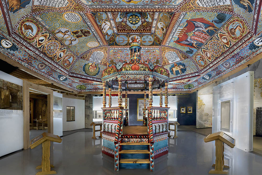 Elaborately painted domed ceiling with intricate patterns and animal motifs above a wooden decorated platform inside a museum interior
