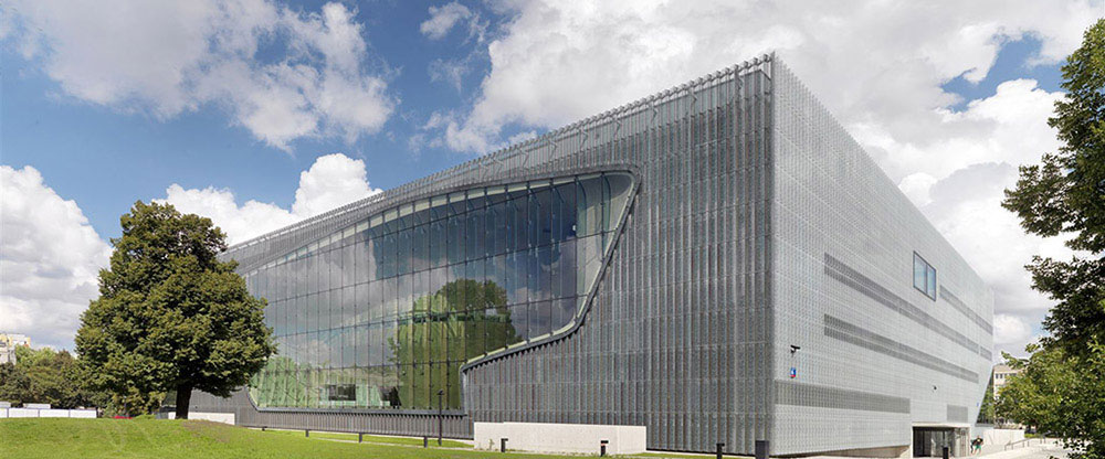 Modern angular building with perforated metal facade and curved glass window, viewed from a grassy park with trees under a bright sky